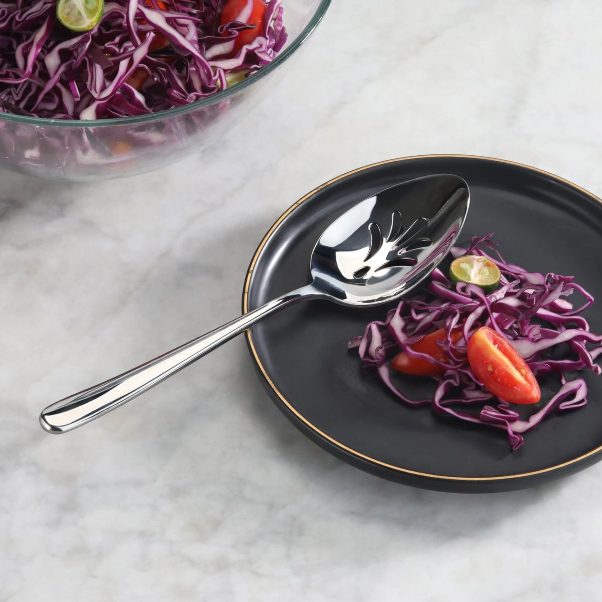 Slotted stainless steel serving spoon with mirror finish on a black plate with red cabbage and tomato slices.