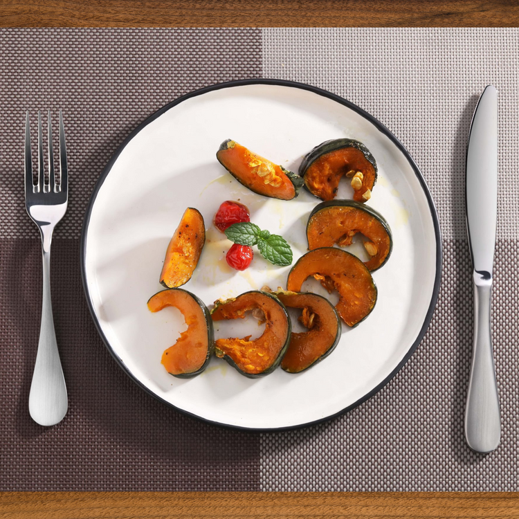 White plate with roasted pumpkin slices garnished with mint and cherry tomatoes, placed between a fork and a stainless steel