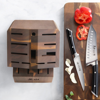 
                    
                      Empty angled acacia wood knife block with 16 slots next to a cutting board with two knives, garlic cloves, and sliced red bel
                    
                  