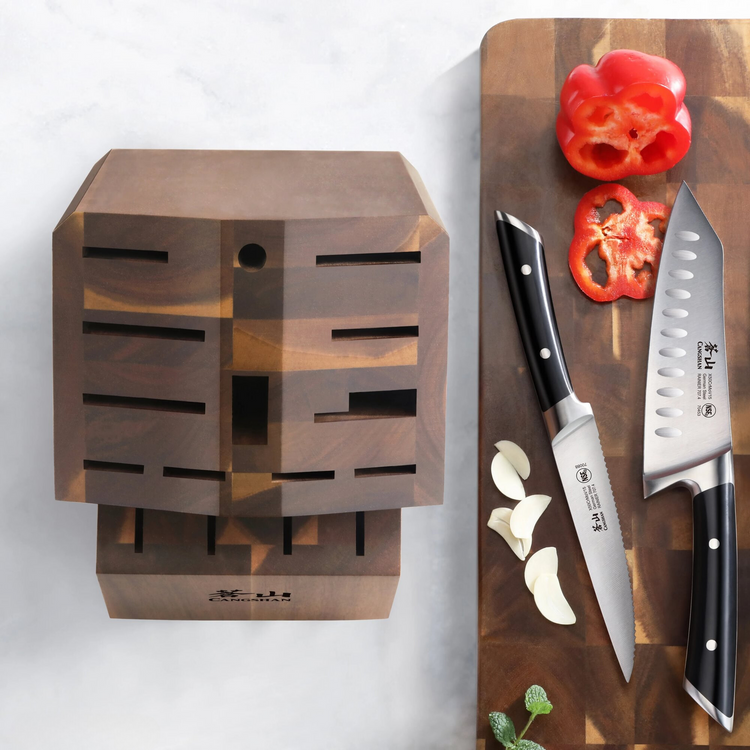 Empty angled acacia wood knife block with 16 slots next to a cutting board with two knives, garlic cloves, and sliced red bel