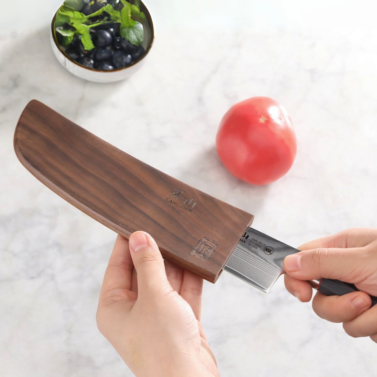 Hand holding a walnut wooden knife sheath while another hand pulls out a kitchen knife, with a tomato and bowl of greens in t