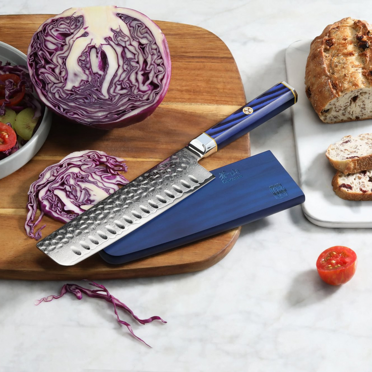 Nakiri knife with hammered blade and blue handle on wooden board with sliced red cabbage, bread, and tomato.