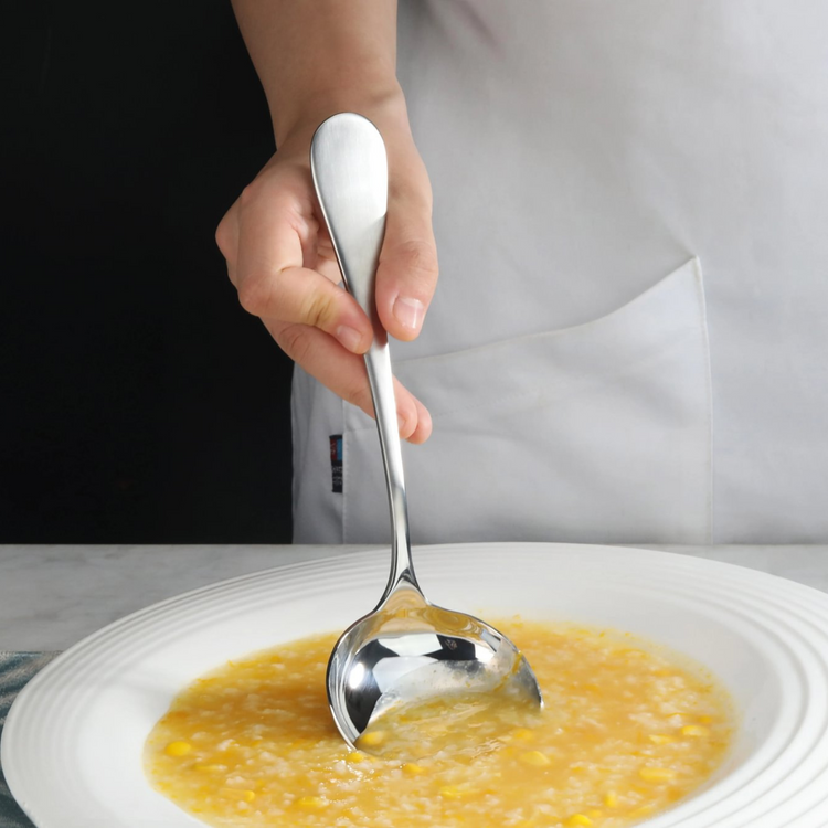 Hand holding a stainless steel soup ladle with a matte finish handle, scooping corn soup from a white bowl.