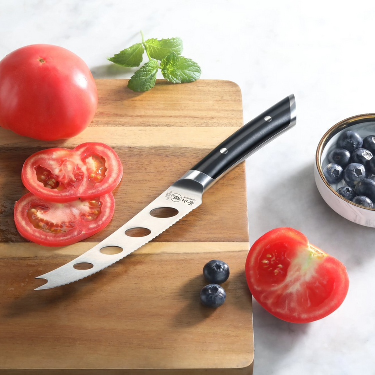 Tomato knife with black handle and holes in blade on wooden cutting board with sliced tomato and blueberries nearby.