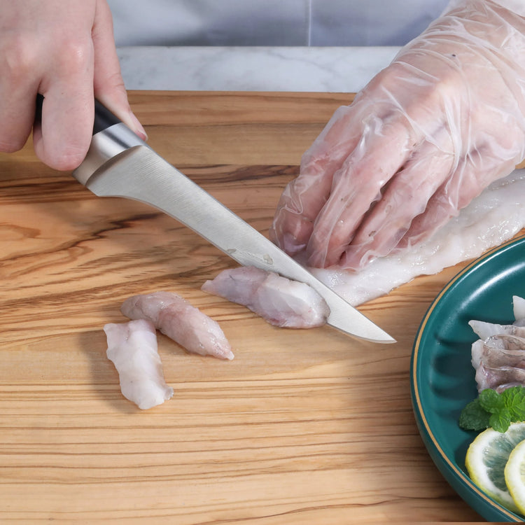 Hand with glove filleting white fish on wooden board using a black-handled fillet knife.