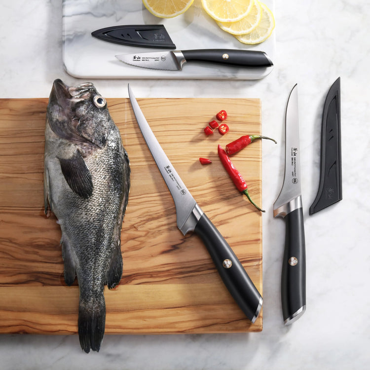 Three black-handled fillet knives with sheaths displayed on a wooden board and marble surface, alongside a whole fish, lemon