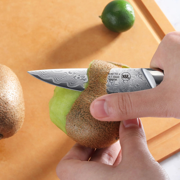 Hand holding a paring knife peeling the skin off a kiwi on a wooden cutting board with a lime nearby.