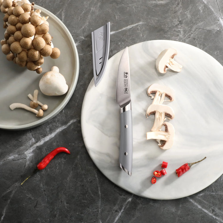 3.5-inch paring knife with gray handle and sheath on a marble board with sliced mushrooms and red chili peppers.