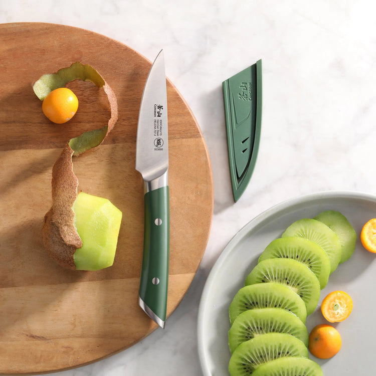 Paring knife with green handle and sheath next to peeled kiwi and sliced kiwi on a wooden board and plate.