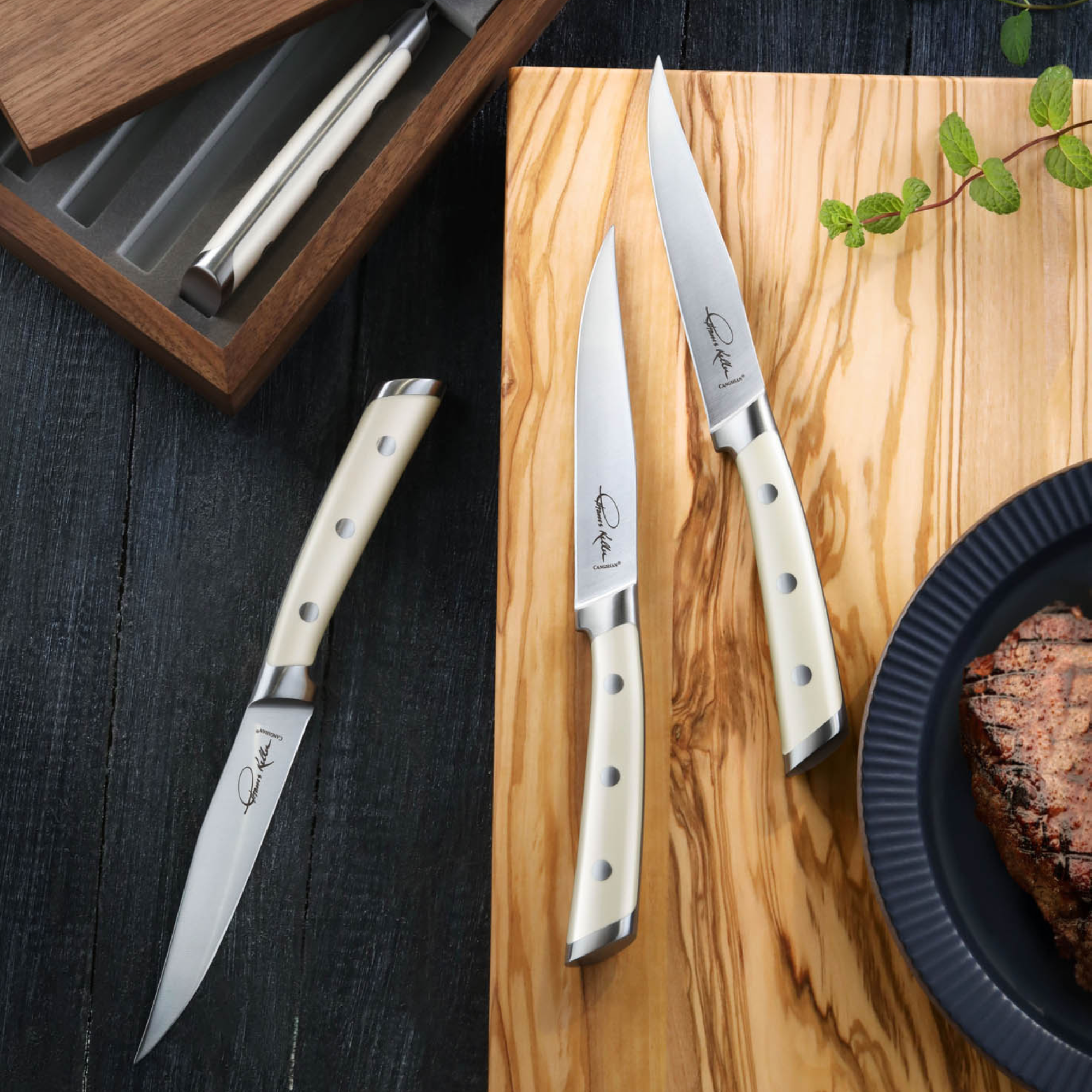 Four white-handled steak knives with straight edges, three on a wooden board and one near a walnut box.