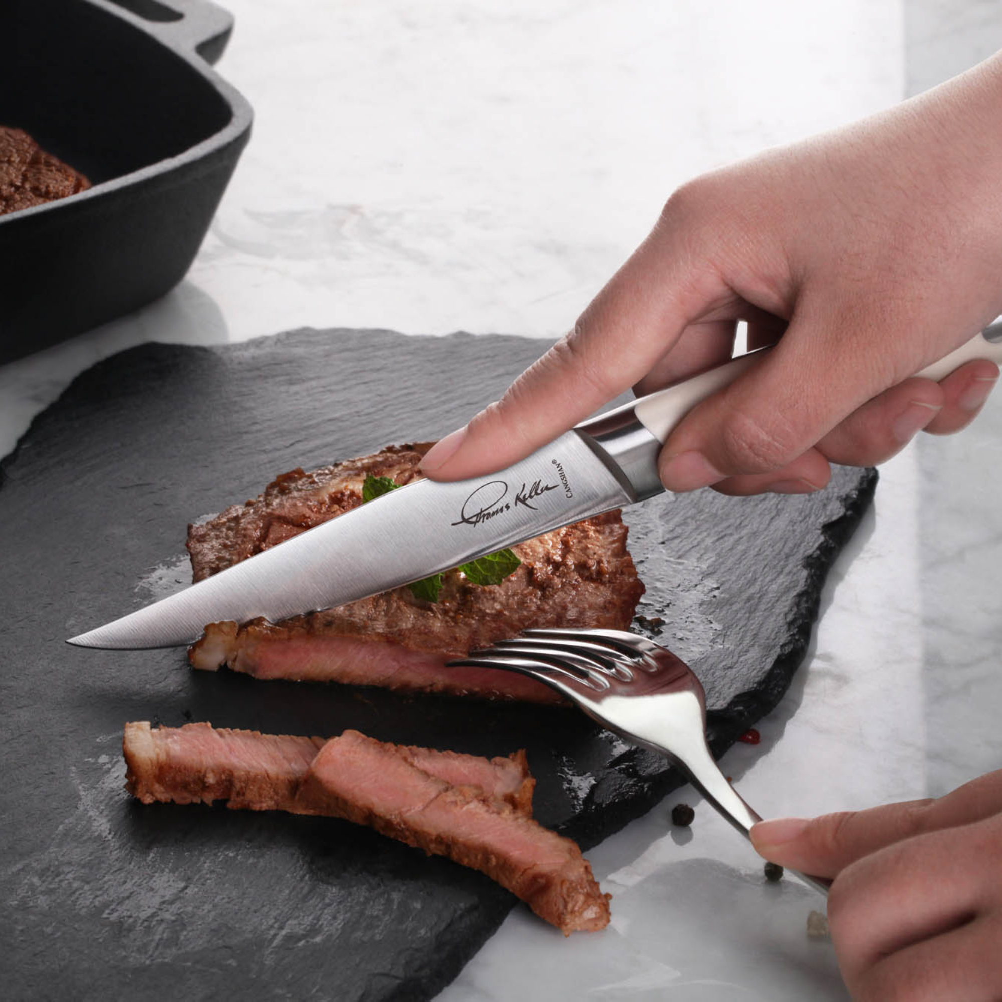 Hand holding a steak knife cutting a cooked steak on a black slate plate, with a fork securing the meat.