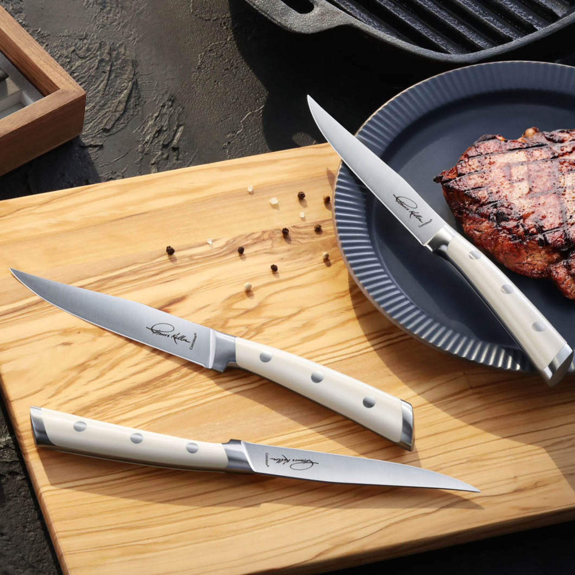 Three white-handled steak knives with straight edges on a wooden cutting board and a blue plate holding a grilled steak.