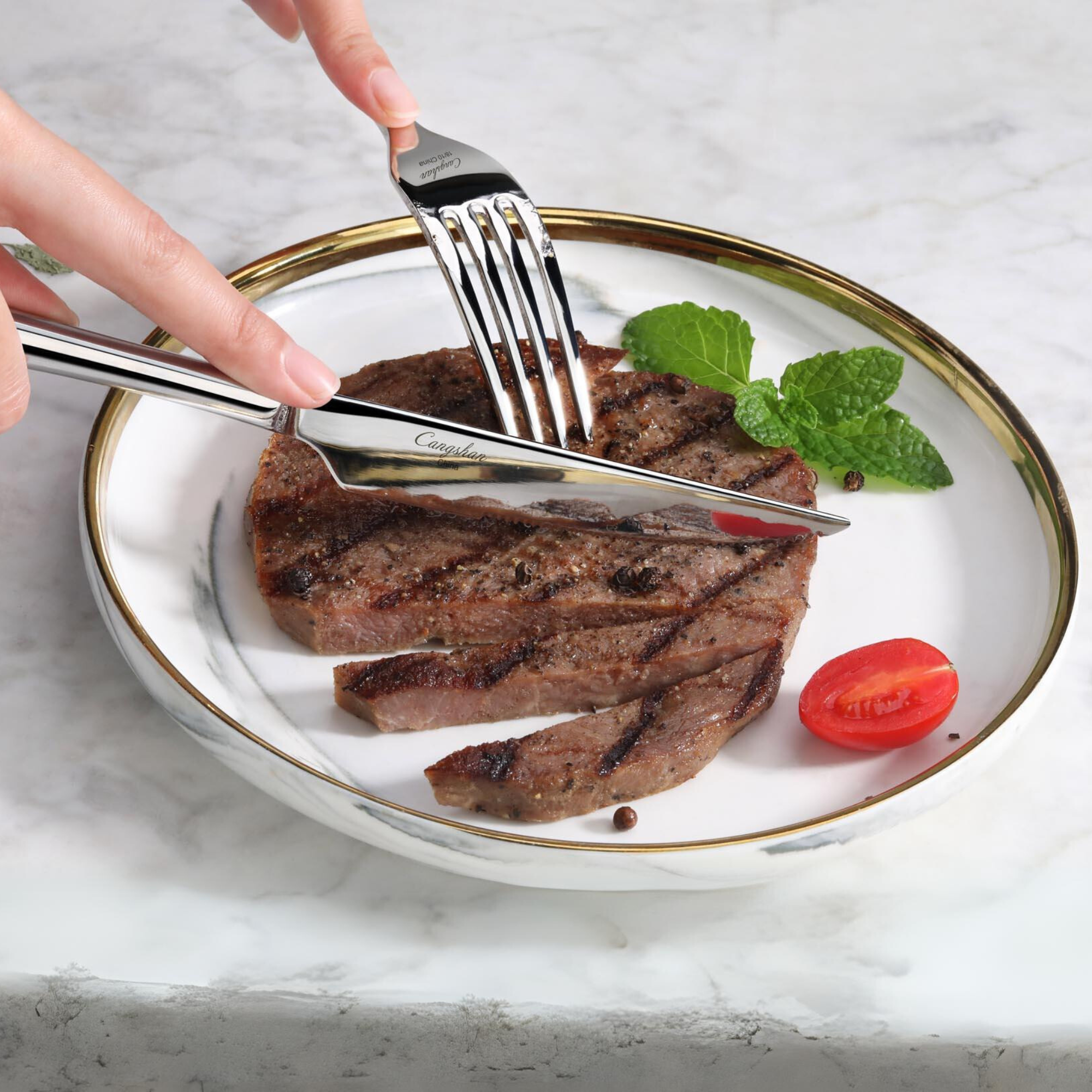 Fork and knife cutting a grilled steak on a white plate with a cherry tomato and mint garnish.