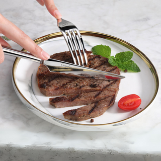 
                    
                      Fork and knife cutting a grilled steak on a white plate with a cherry tomato and mint garnish.
                    
                  