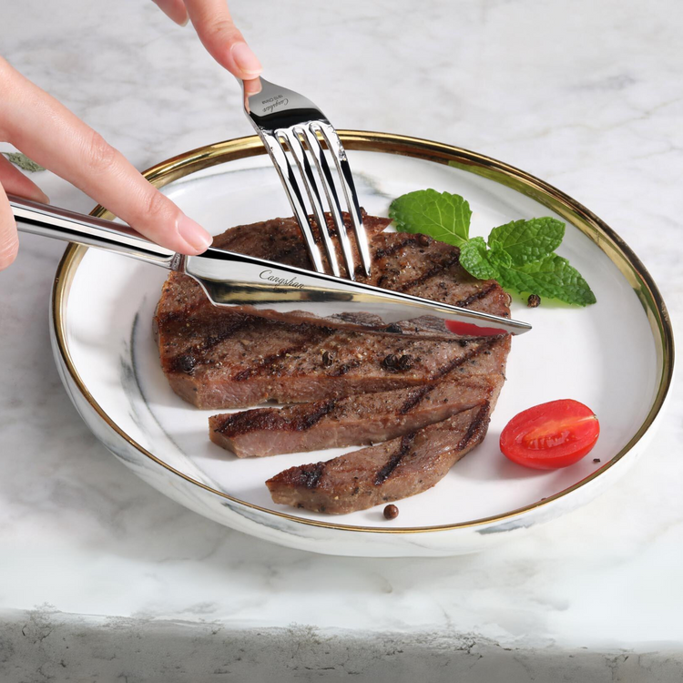 Fork and knife cutting a grilled steak on a white plate with a cherry tomato and mint garnish.