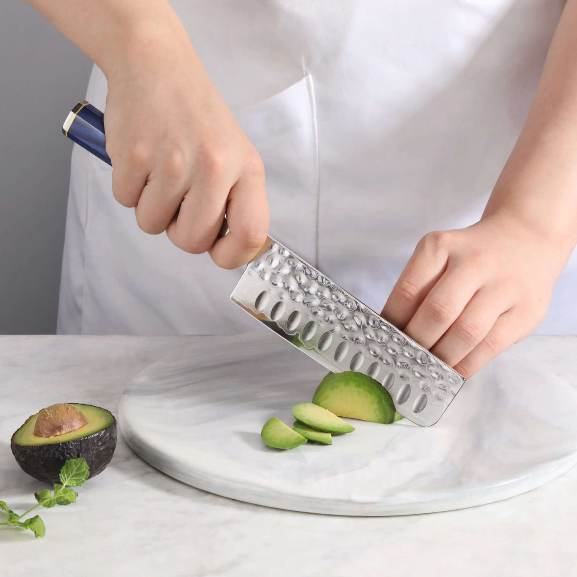 Hand holding a Nakiri knife slicing an avocado on a round marble cutting board.