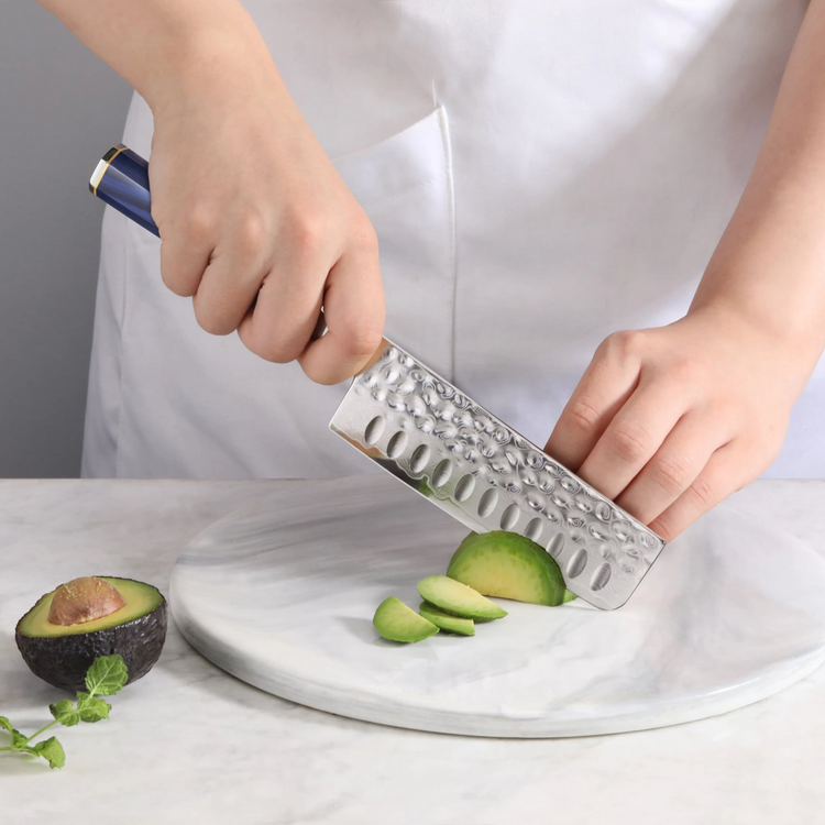 Hand holding a Nakiri knife slicing an avocado on a round marble cutting board.