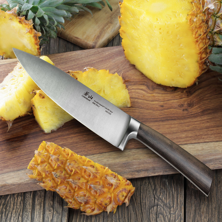 Chef knife with dark wooden handle slicing fresh pineapple on a wooden cutting board.