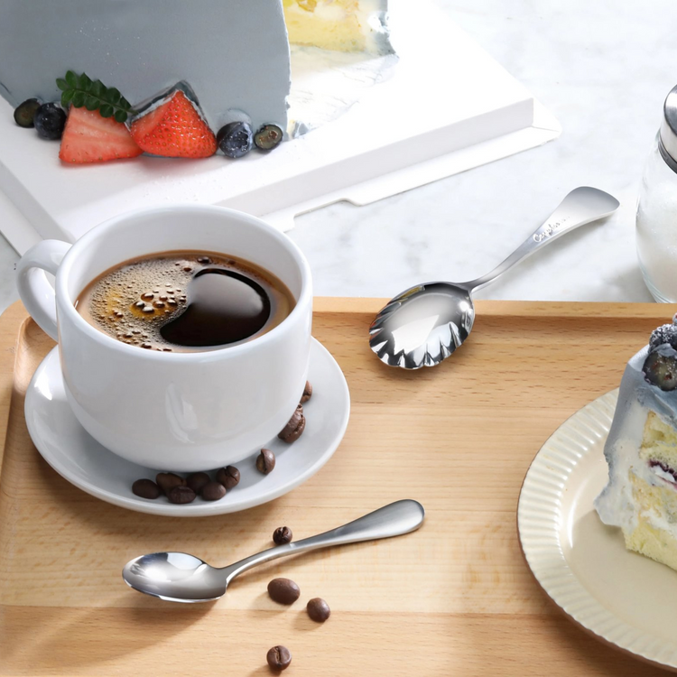 Stainless steel espresso spoon with matte finish handles on a wooden tray next to a cup of coffee and slices of cake.