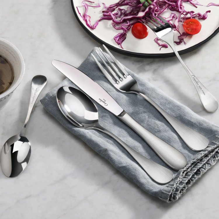 Stainless steel dinner knife, fork, and spoon with matte finish handles on a gray napkin next to a plate with salad.