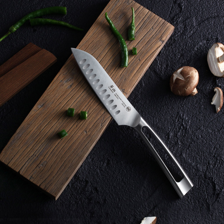 Santoku knife with a stainless steel blade and handle on a wooden board, surrounded by green chili pieces and mushrooms.