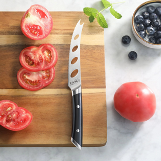 
                    
                      Tomato knife with black handle and holes in blade on wooden cutting board with sliced tomato and blueberries nearby.
                    
                  