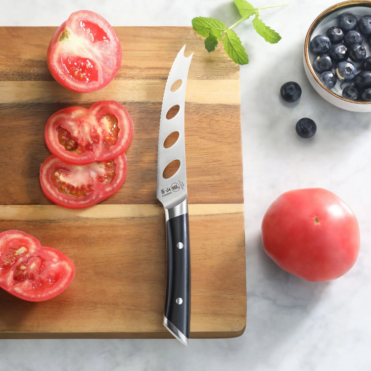 Tomato knife with black handle and holes in blade on wooden cutting board with sliced tomato and blueberries nearby.