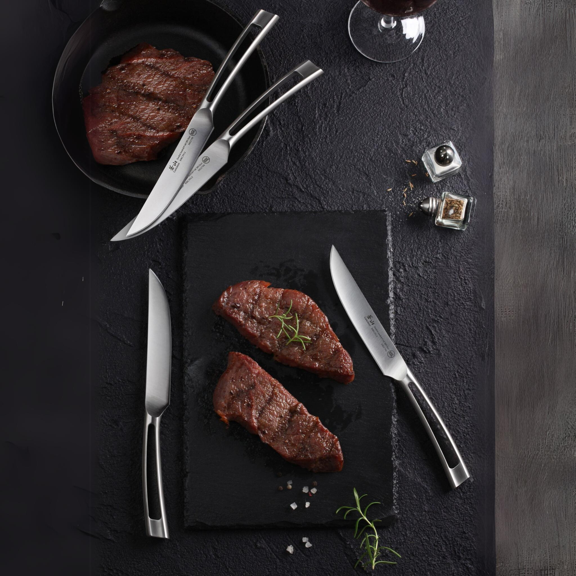 Four stainless steel steak knives arranged around two cooked steak pieces on a dark slate surface with salt, pepper, and rose