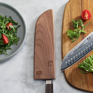 
                    
                      Walnut magnetic knife sheath covering a chef's knife next to a cutting board with chopped greens and tomato.
                    
                  