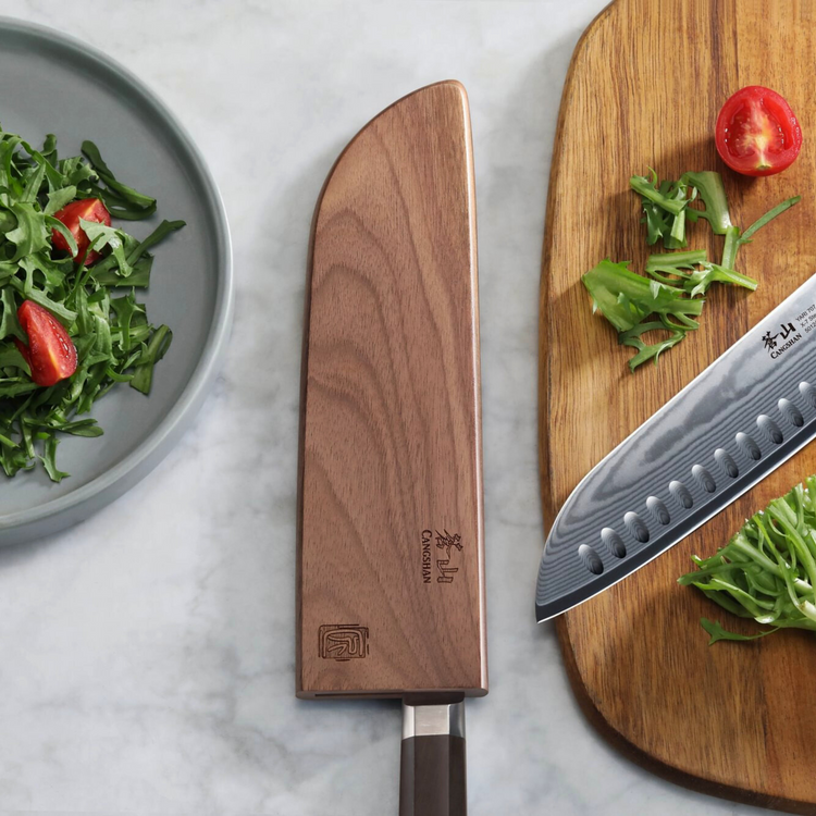 Walnut magnetic knife sheath covering a chef's knife next to a cutting board with chopped greens and tomato.