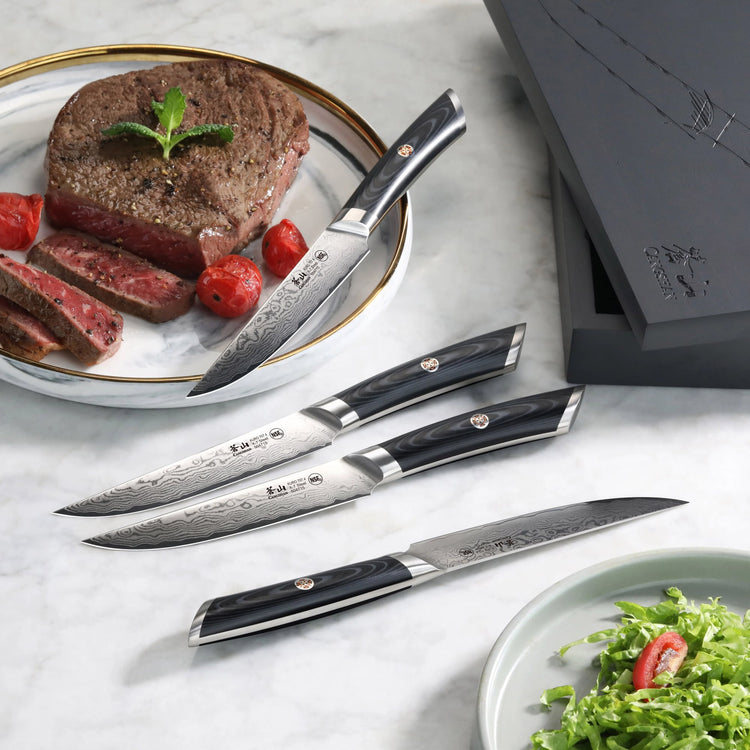 Four steak knives with patterned blades and dark handles displayed next to sliced steak on a plate and a black storage box.