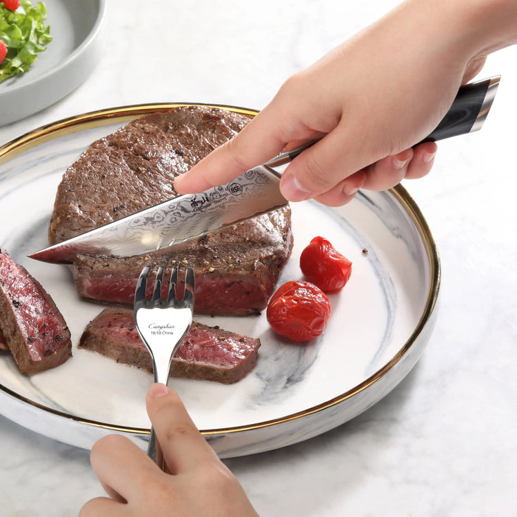 Hand cutting medium-rare steak on white plate with gold rim using patterned steak knife and fork.