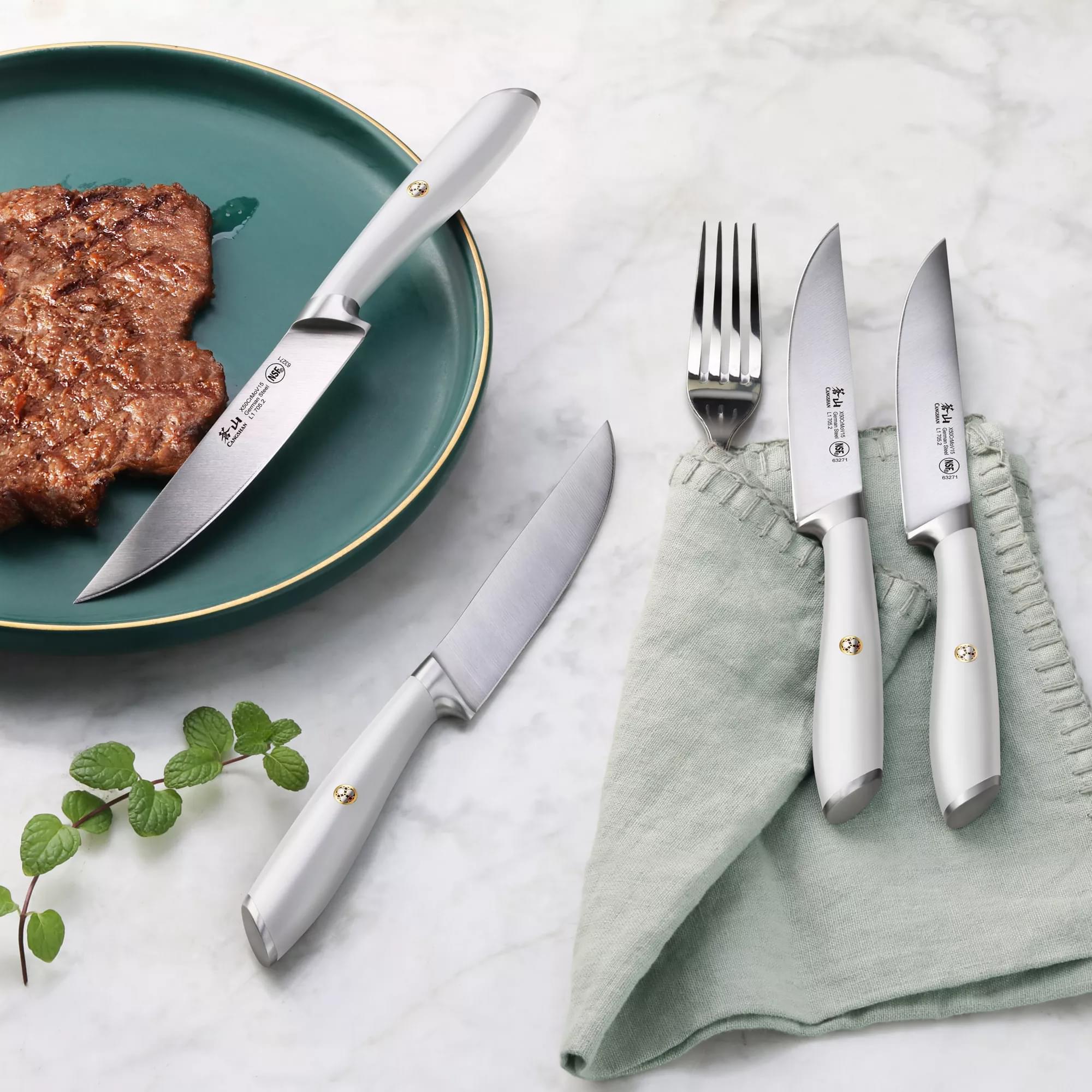 Four stainless steel steak knives with white handles, one fork, a cooked steak on a green plate, and a green napkin on a marb