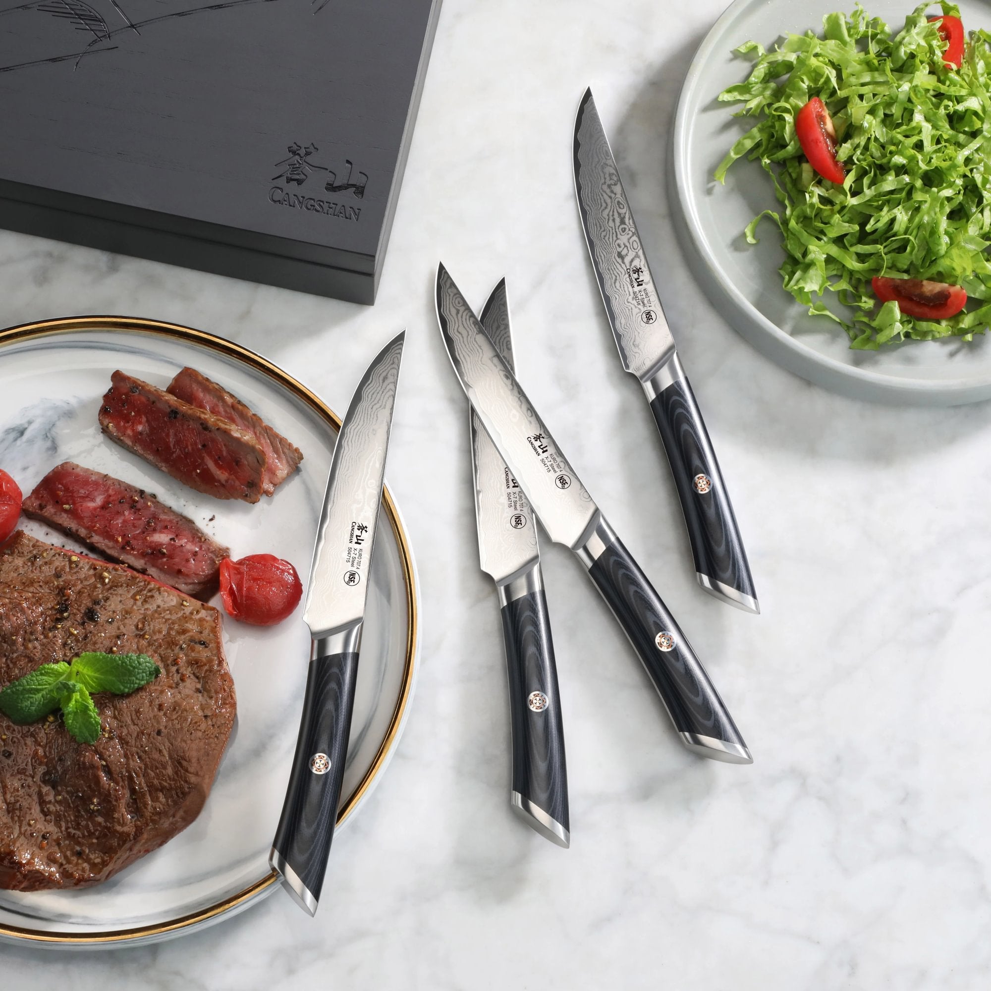 Four steak knives with black handles and patterned blades next to a plate with cooked steak and a salad bowl.