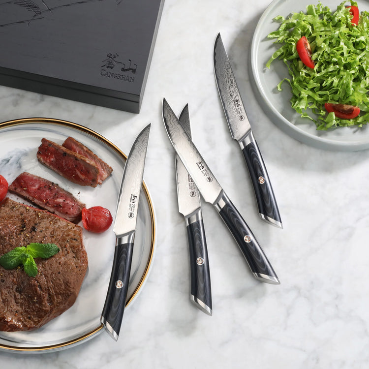 Four steak knives with black handles and patterned blades next to a plate with cooked steak and a salad bowl.