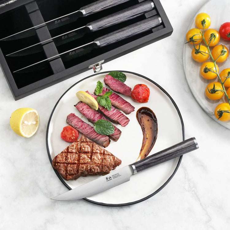 Four steak knives with pakkawood handles in a wooden box, one knife cutting sliced steak on a plate with grilled vegetables.