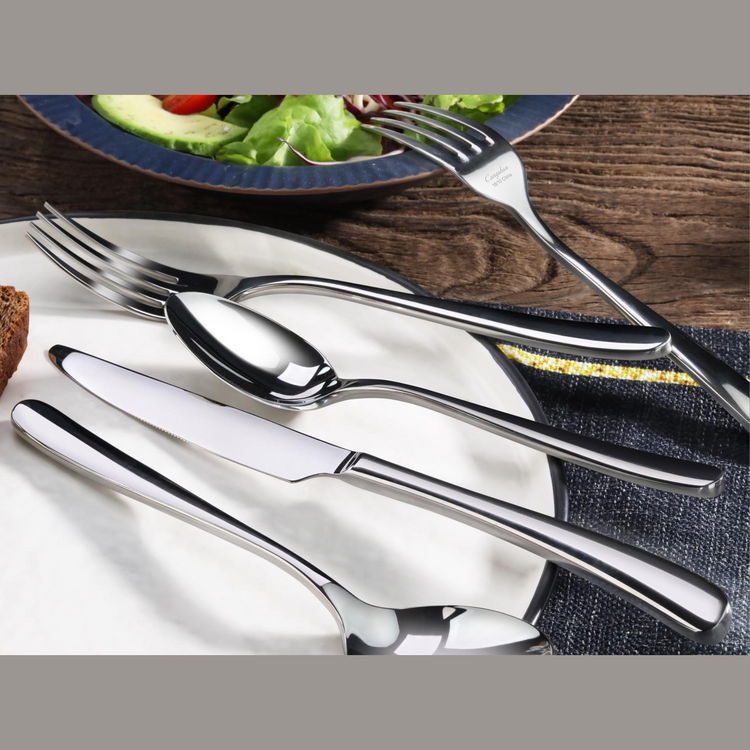 Stainless steel fork, spoon, and knife arranged on a white plate with a salad plate in the background.
