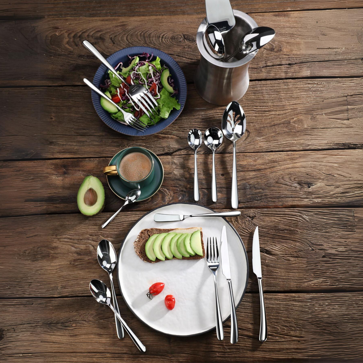 Stainless steel flatware set displayed with a salad bowl, avocado toast on a plate, coffee cup, and wooden table.