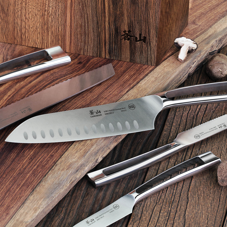 Six stainless steel knives with hollow edges and polished handles arranged on a wooden surface near a walnut knife block.