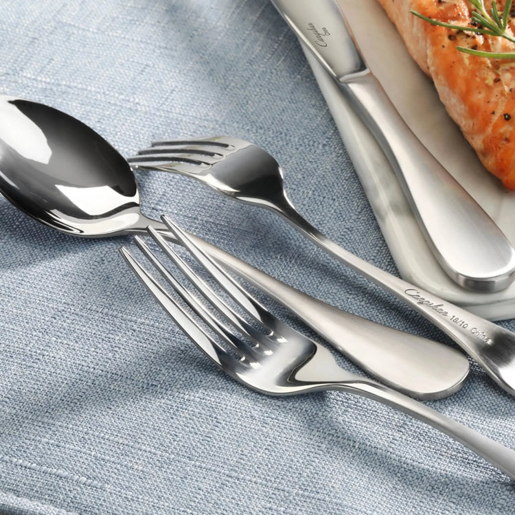 Stainless steel flatware with matte finish handles arranged on a blue cloth next to a plate with cooked salmon.