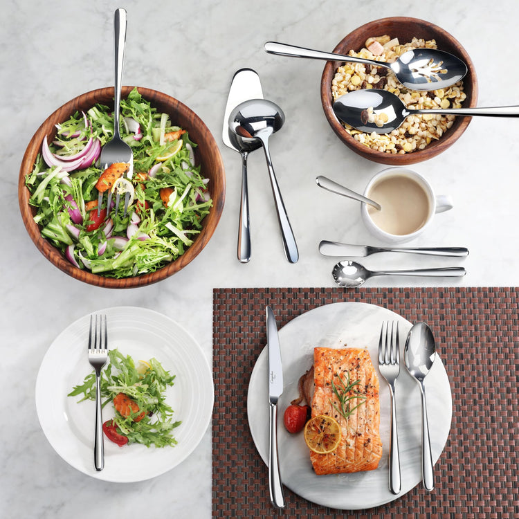 Wooden bowls with salad and popcorn, stainless steel serving utensils, and plates with salad and cooked salmon on a marble su