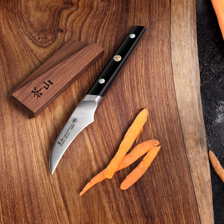 Japanese-style paring knife with black handle on wooden cutting board next to peeled carrot pieces and wooden sheath.