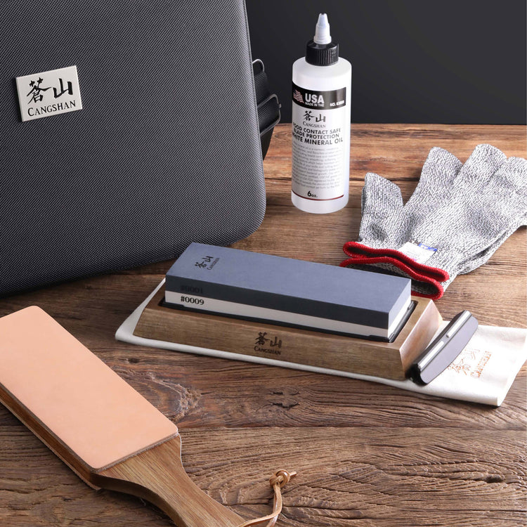 Dual-sided whetstone on wooden base with sharpening oil, cut-resistant glove, leather strop, and carrying case on wooden surf