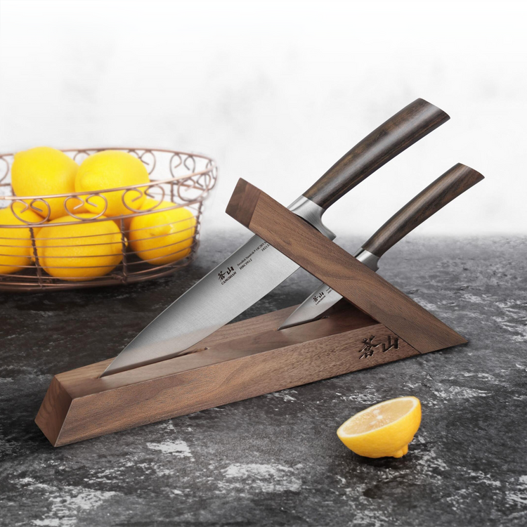Two knives with dark wooden handles in a slanted walnut knife block on a dark countertop, with lemons nearby.