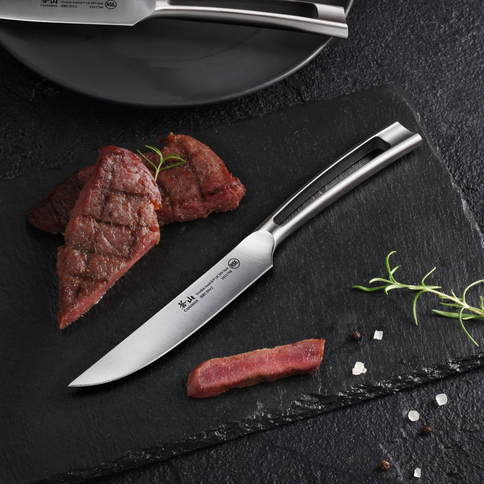 Steak knife with stainless steel handle on slate board, sliced grilled steak, rosemary sprig, salt, and peppercorns.
