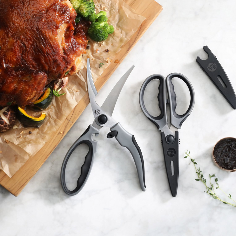 Gray heavy-duty kitchen shears set with serrated blades and protective guards next to cooked meat and vegetables.