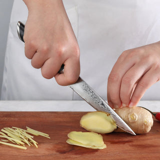 
                    
                      Hand holding serrated utility knife slicing ginger on wooden cutting board with thin ginger strips nearby.
                    
                  