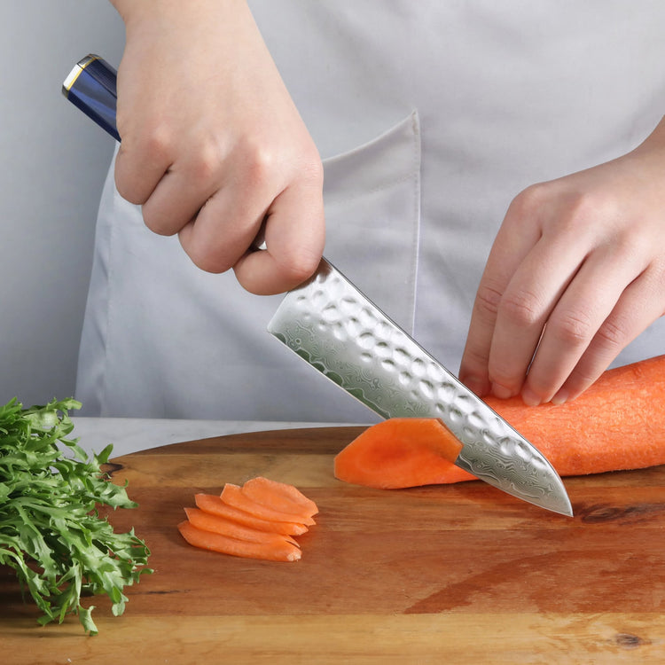 Hand holding a blue-handled Damascus steel utility knife slicing a carrot on a wooden cutting board with greens nearby.