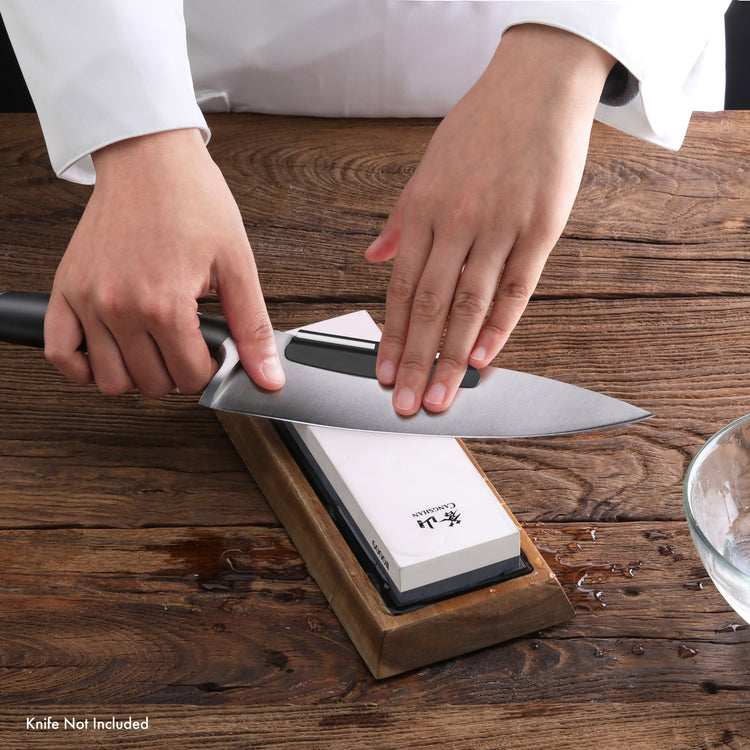 Hands sharpening a chef's knife on a dual-sided whetstone with wooden base on a wooden surface.
