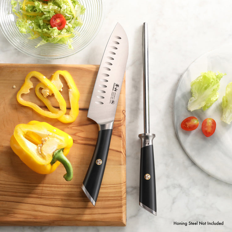 Santoku knife and honing steel with black handles on a wooden board next to sliced yellow bell pepper and salad ingredients.